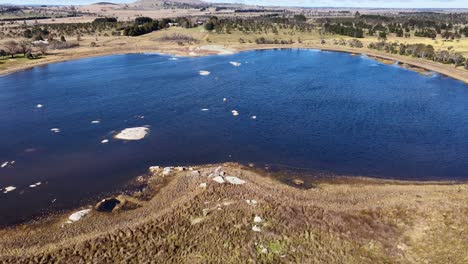 drone camera glides above a blue pond bordered by grassy fields, scattered trees, and rocks under bright daylight, capturing a tranquil rural landscape