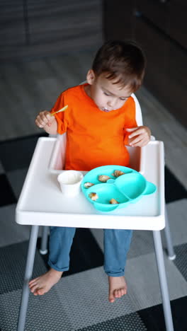 three-year-old toddler boy sits at high chair. cheerful baby eating himself at home. top view. vertical video.
