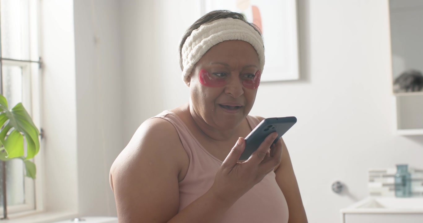Donna afroamericana con gel rossi per gli occhi che guarda in basso, leggendo messaggi sul telefono, toccando vicino alla toeletta.