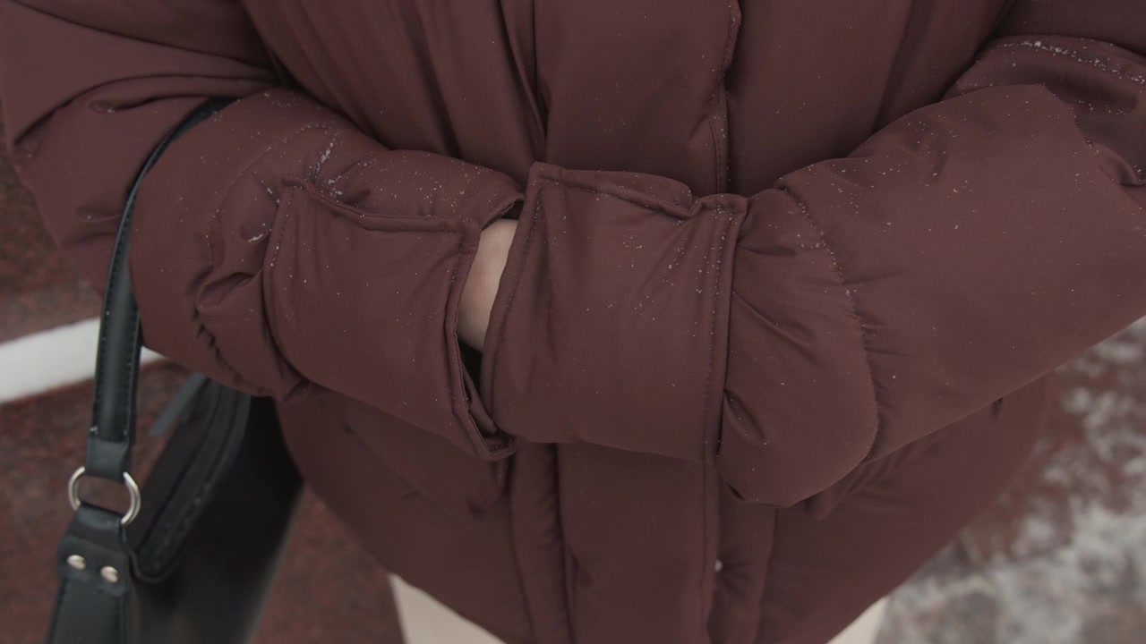 Close-up of person in brown puffer jacket on snowy sidewalk, hands pulling front closed with tucked posture, showing subtle warming movement, protective feel, winter streetwear detail and calm