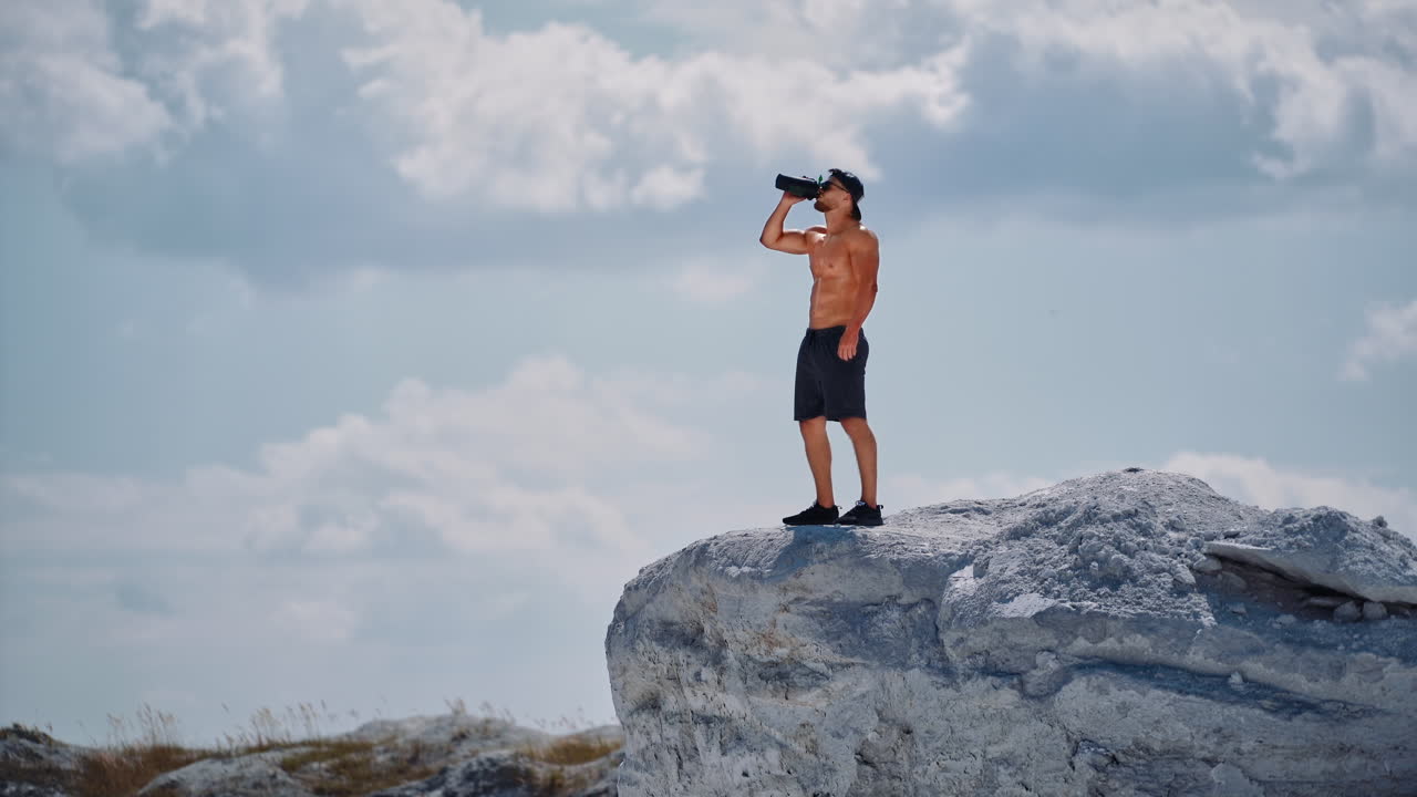 Muscular male in shorts only drinking water after hard workout outdoors. Bodybuilder standing on white rock with flask on the sky background.
