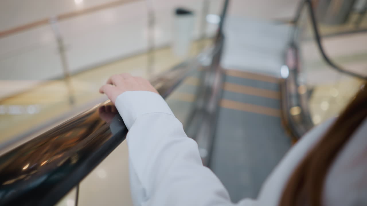 primer plano de una mujer con uñas pulidas negras tocando la barandilla de la escalera mecánica mientras desciende, lleva una blusa blanca con mangas largas, el fondo borroso muestra el interior moderno de un centro comercial