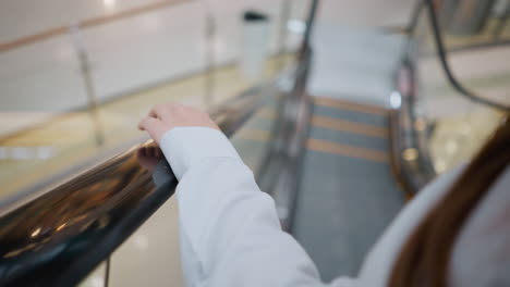 close-up of lady with black polished nails tapping escalator handrail while descending, she wears a white blouse with long sleeves, blurred background showcases modern shopping mall interior