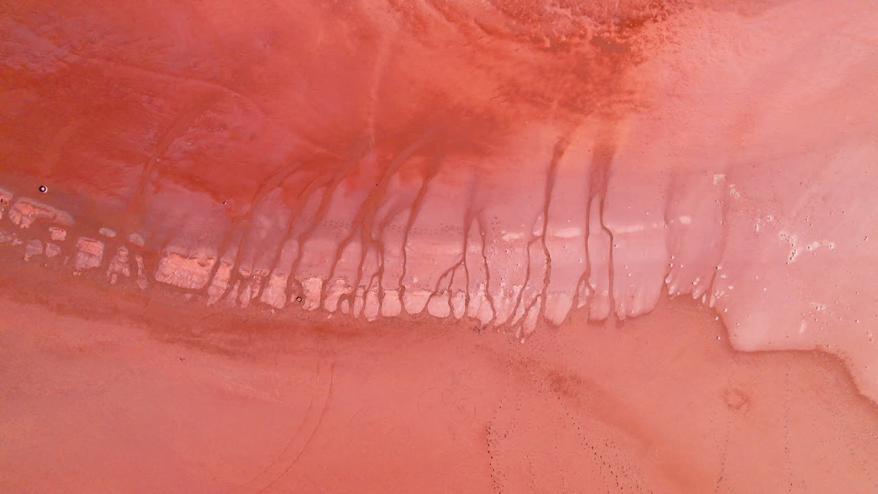 Top-down aerial shot of a dried-out salt lake with soft pink tones still visible in the surface. The cracked textures, pastel colours, and wide open terrain create a unique abstract landscape