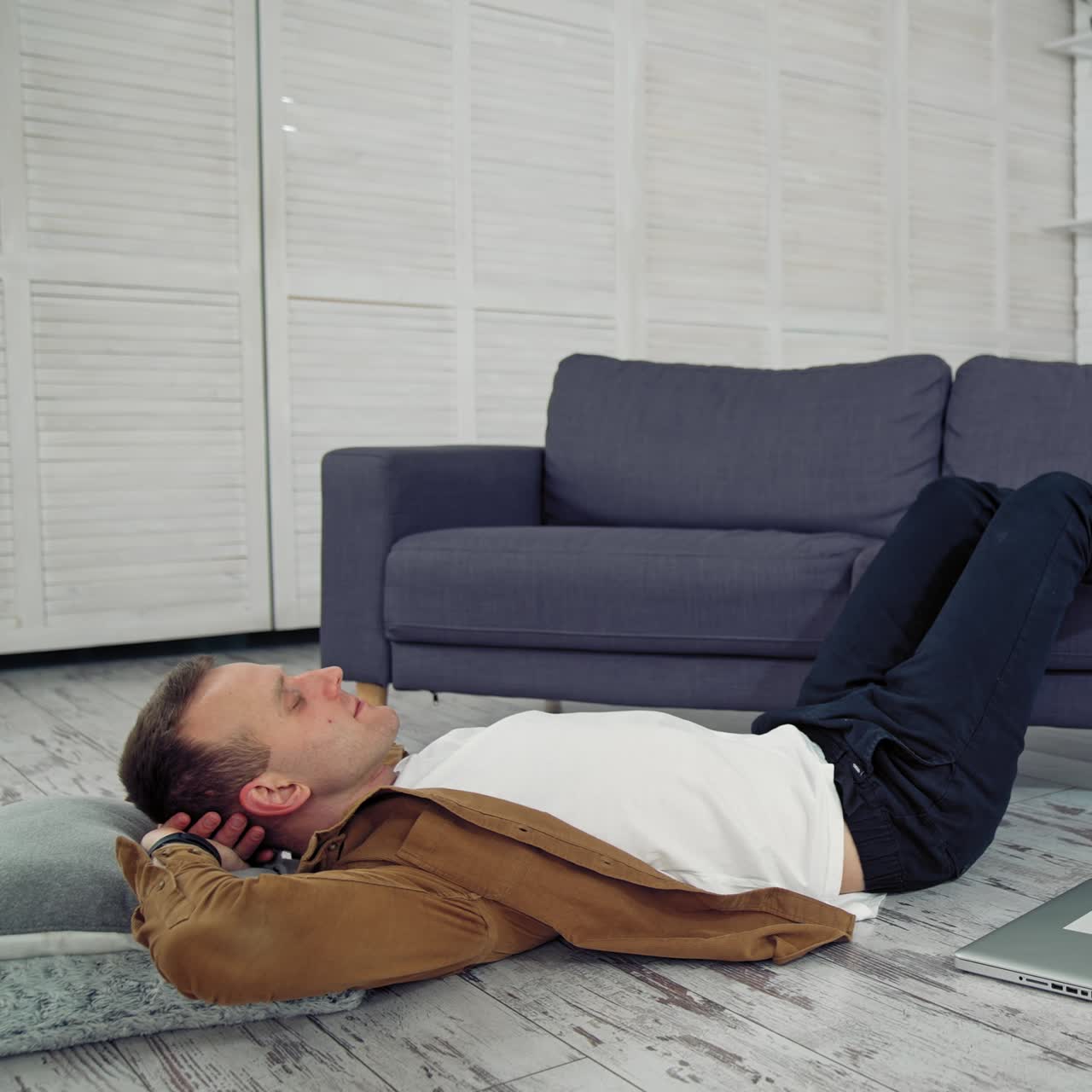 Tired freelance worker rests on floor. Young guy sleeping on the kitchen floor with his legs on sofa. Laptop lying on the floor. Remote job. Quarantine. Lockdown concept