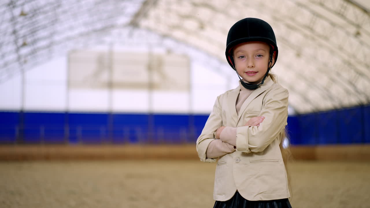 Young confident horse rider in black jockey helmet stands holding hands folded on her chest. Girl touché her hat looking at camera. Close up. Blurred backdrop.