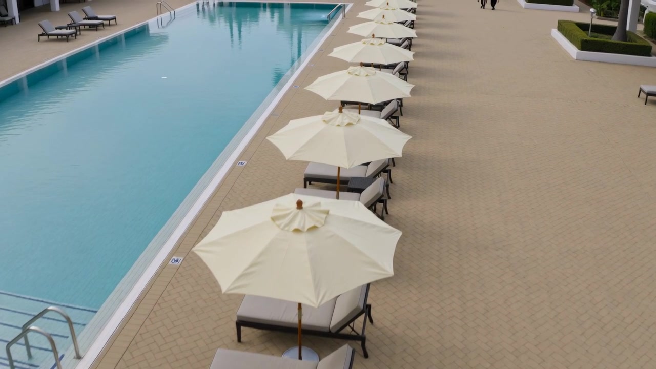 Starting camera glide following long hotel pool deck, revealing umbrellas, chairs, distant walkers