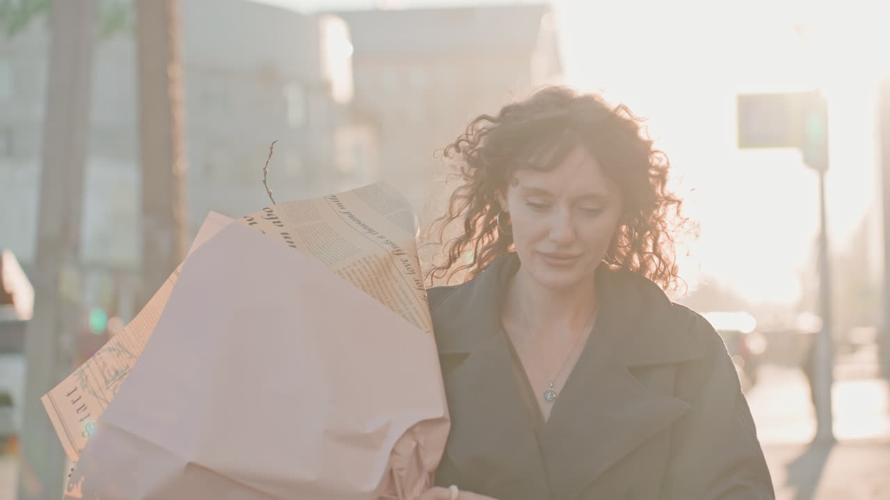 Clienta de floristería caminando por una calle de la ciudad con un ramo a contraluz del atardecer, mujer caucásica cruzando una intersección, el viento revolviendo su cabello rizado, ambiente espontáneo de una viajera con destello de lente y resplandor urbano.