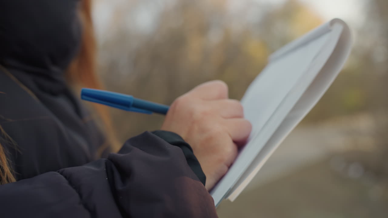 Mezza figura di persona che scrive su un quaderno con una penna blu, giacca isolante e manica visibile, viale alberato con bokeh morbido, annotazioni deliberate e giri di pagina costanti, atmosfera documentaristica e pratica.