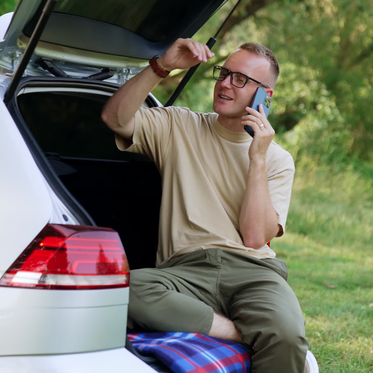 Relaxed Caucasian man sitting in the trunk of his car speaks on the phone. Working remote, business conversation in nature