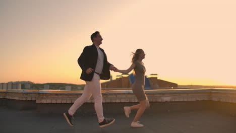 young couple holding hands walking woman leading boyfriend the roof of the building at sunset pov travel concept. carefree free lovers run on the roof laughing and smiling