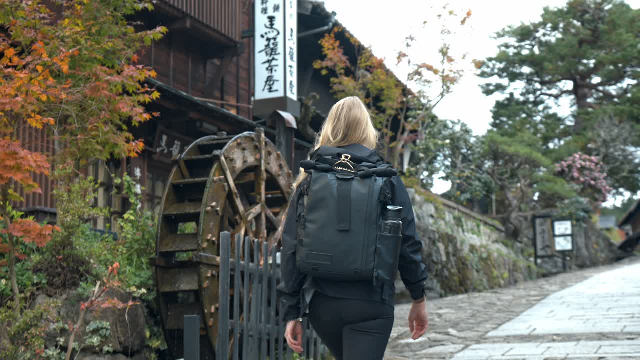 A woman walking along the historic Nakasendo Trail, passing by a traditional water mill in the charming village of Magome, Japan.