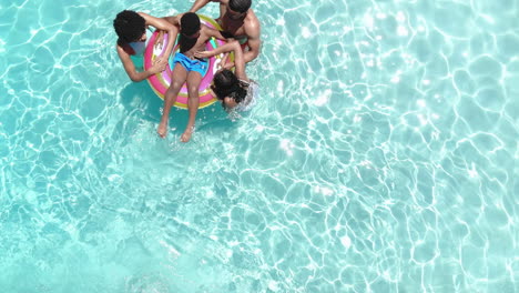 Aerial-of-african-american-parents,-son-and-daughter-playing-on-inflatable-in-sunny-pool,-copy-space