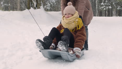 father and mother help their daughter to ride on sled in snowy forest