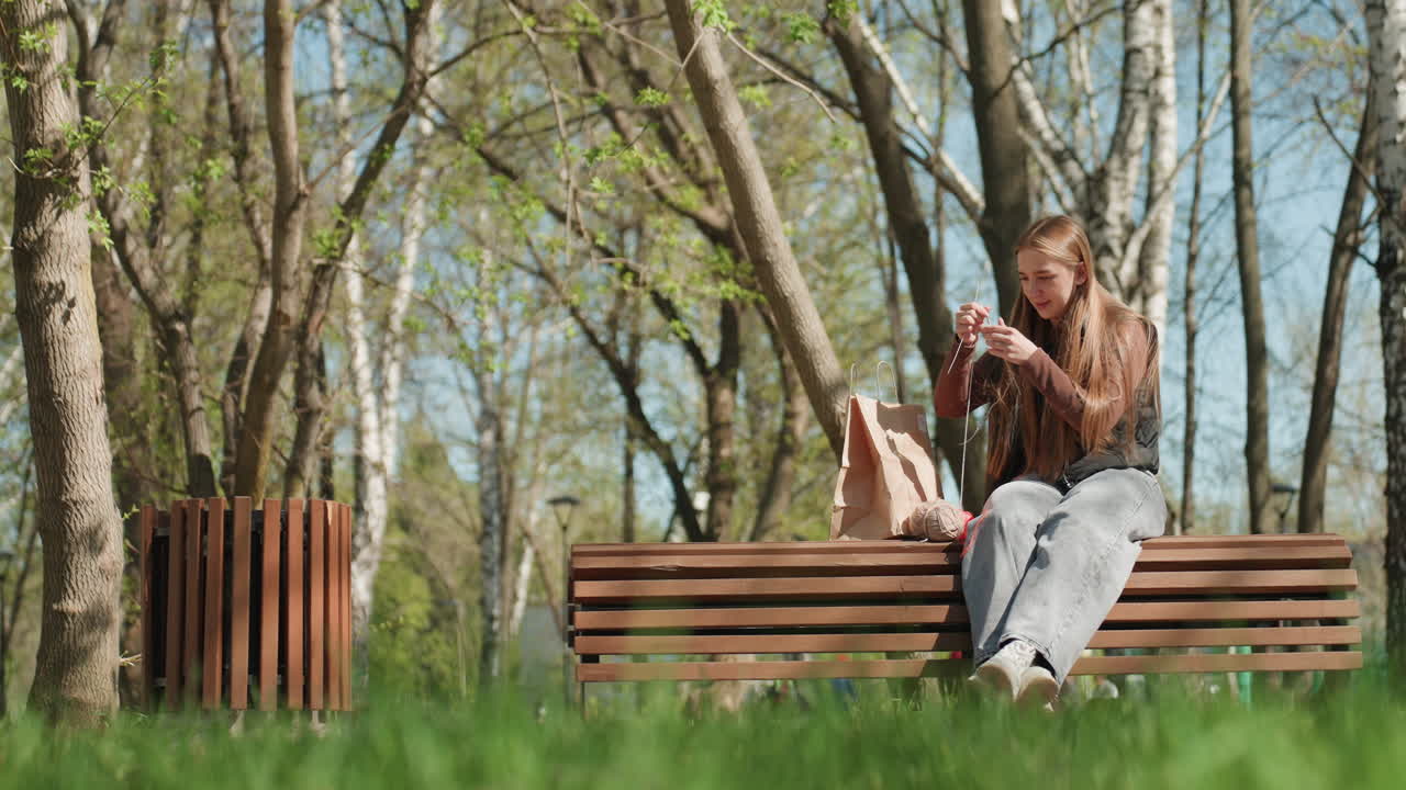 자작나무 숲 벤치에 앉아 스웨터를 수선하는 백인 여행객, 근처에 백팩을 둔 채 집중한 손으로 실을 조심스럽게 꿰매고 있어요. 조용히 옷을 수선하는 장면, 차분한 집중, 봄 햇살.