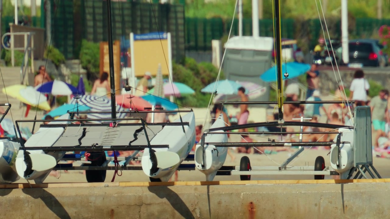 Catamarans au repos sur un quai en béton, surplombant une plage bondée avec des gens profitant des activités estivales