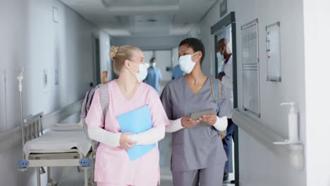 Happy-diverse-female-doctors-wearing-face-masks-talking-and-walking-in-corridor,-slow-motion