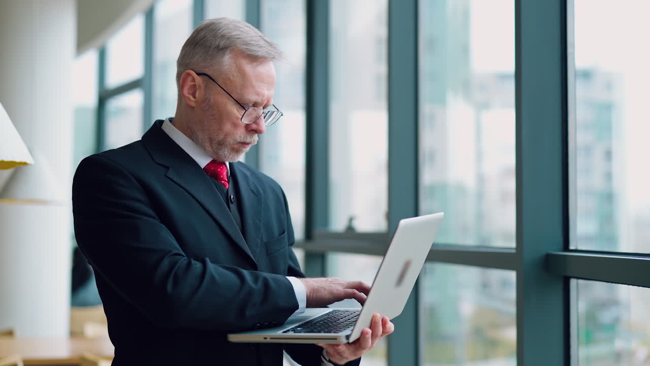 Seriøs forretningsmand med bærbar computer i hænderne tænker på fremtidigt møde og tjekker præsentation, mens han står ved vinduet med panoramaudsigt over byen. Video fra siden.