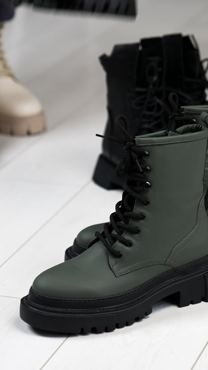 Two pairs of black and grey boots on tractor soles on the floor. Woman in white boots comes up bringing beige and black footwear. Vertical video