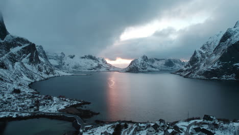 vuelo de avión no tripulado sobre reine, islas lofoten, noruega durante la puesta de sol con el sol asomando a través de nubes oscuras, montañas nevadas y una escena oceánica de mal humor