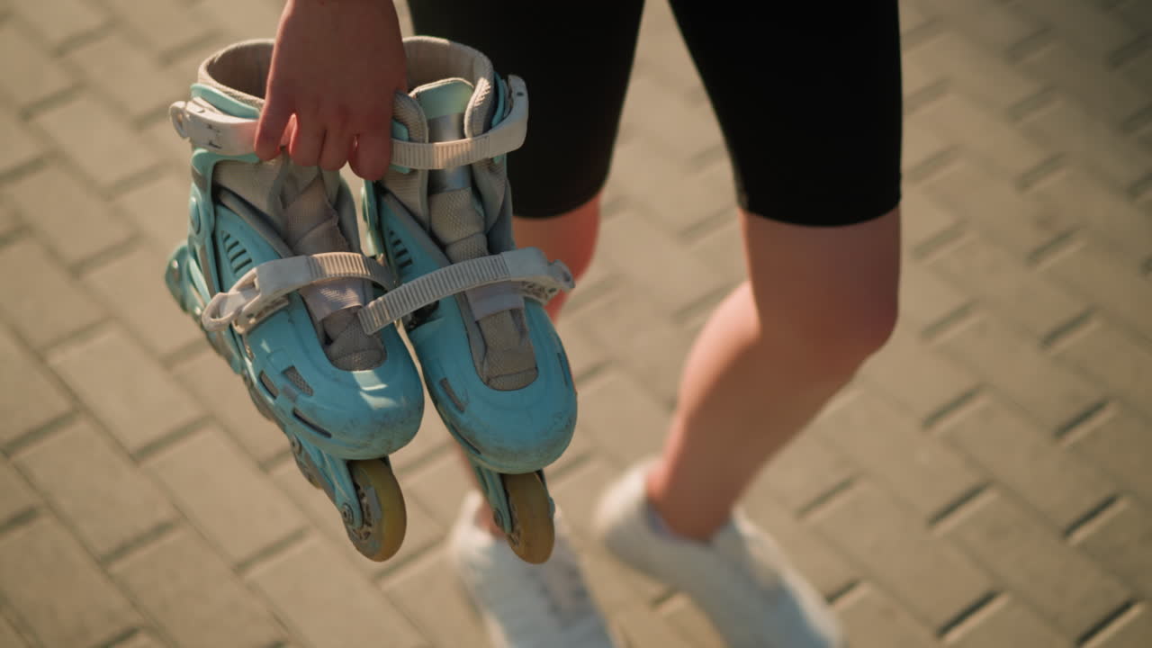 Young lady wearing black tights walking outdoors holding cyan roller skates in her right hand, visible details include a black wristband and an interlocked path background