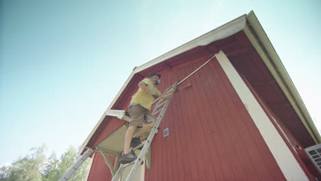 Man-climbs-ladder-with-long-brush-and-removes-peeling-red-paint-from-wooden-home