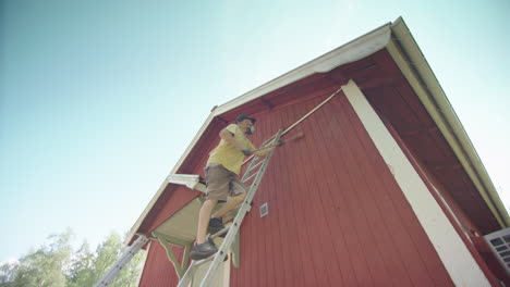 El-Hombre-Sube-La-Escalera-Con-Un-Cepillo-Largo-Y-Quita-La-Pintura-Roja-Pelada-De-La-Casa-De-Madera