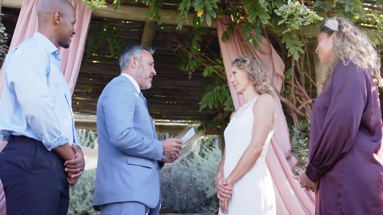 Coppia in piedi sotto un pergolato con l'uomo in abito blu che inizia a leggere le promesse stampate, la donna che ascolta