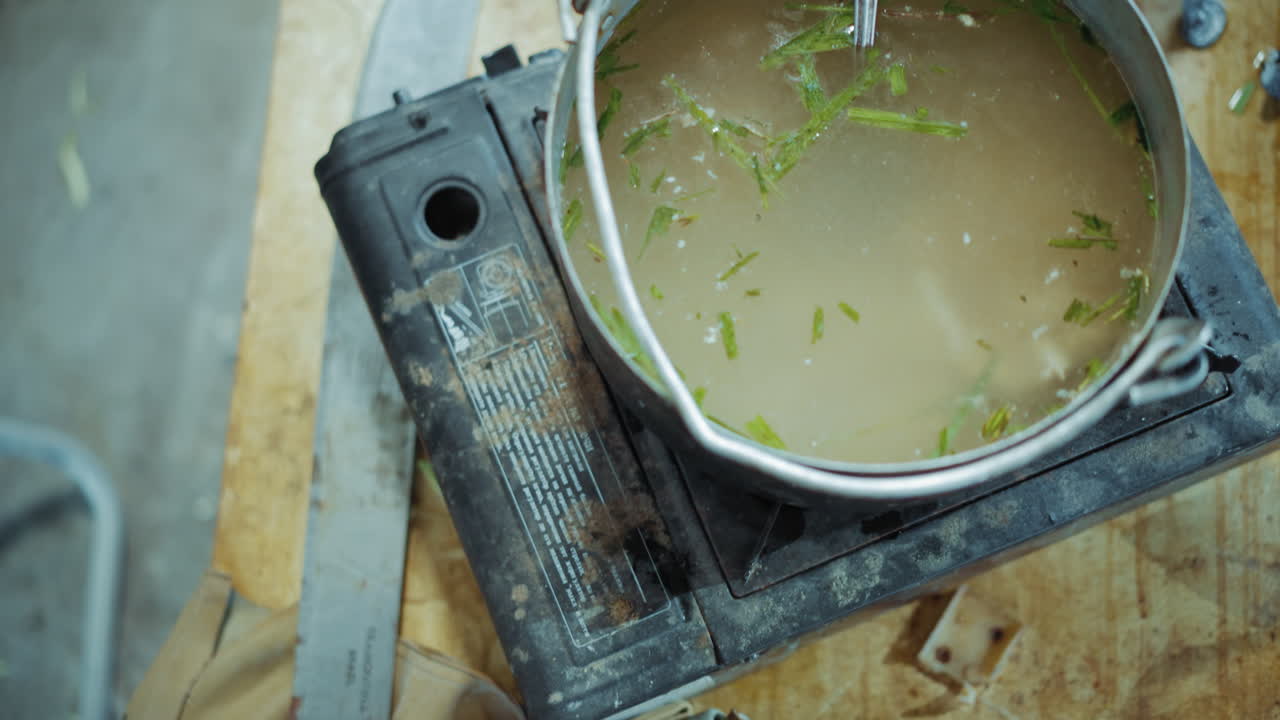 Close up of damaged gas mask placed on wooden table near sharp kitchen knife, old portable stove, and cooking pot, highlighting survival atmosphere