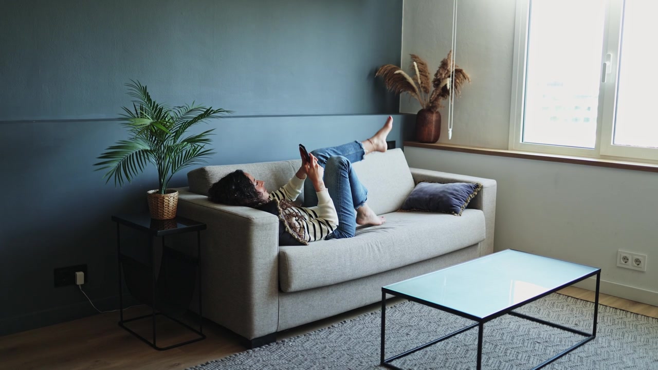 Ung kvinde slapper af på sofaen og bruger smartphone derhjemme