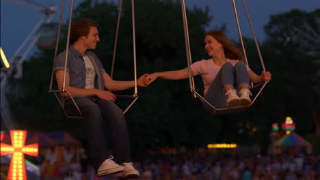 young couple holding hands on a swing ride at a night amusement park