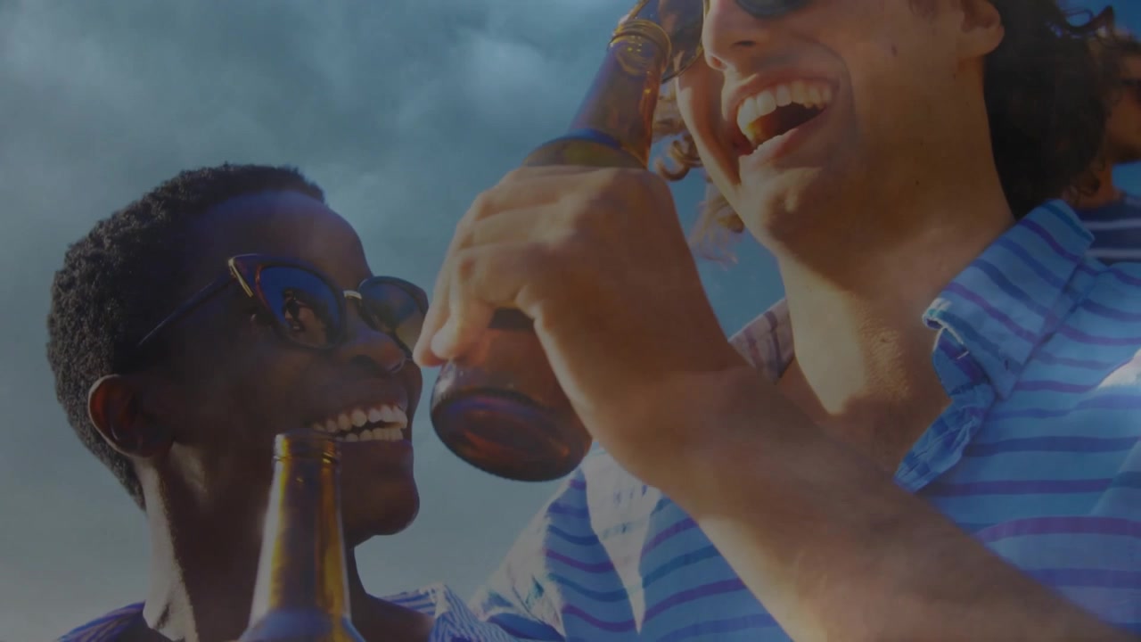 Two friends raising amber bottles toward low camera, clinking then drinking to celebrate sunlight