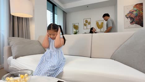 a young girl covers her ears while parents argue in a modern living room. bright lighting and tense atmosphere