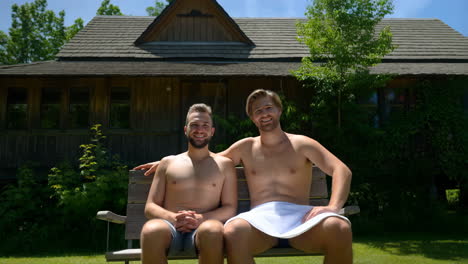 two happy shirtless men sitting on a bench outdoors at a lodge