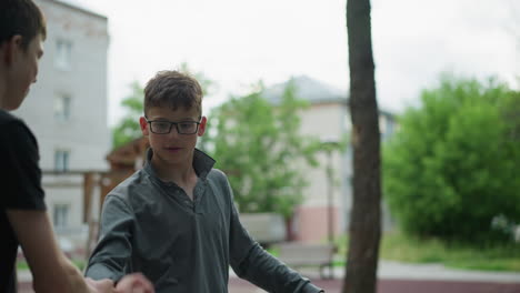 two brothers on bicycles interact playfully, one brother in a grey top reaches out to chop knuckle hands with the other, both smiling in a cheerful moment, with blur background of building