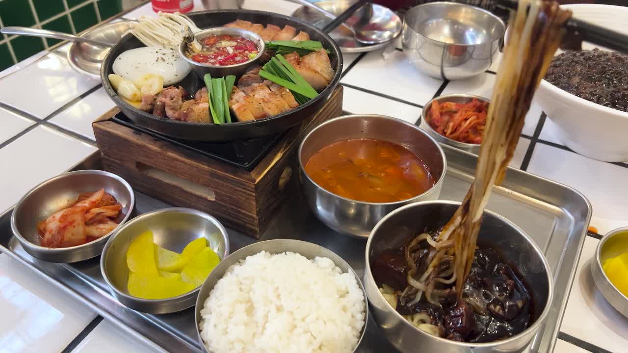 Chopsticks mix and lift jajangmyeon noodles in a vibrant Korean meal setting with grilled pork, rice, kimchi, and side dishes under bright lighting
