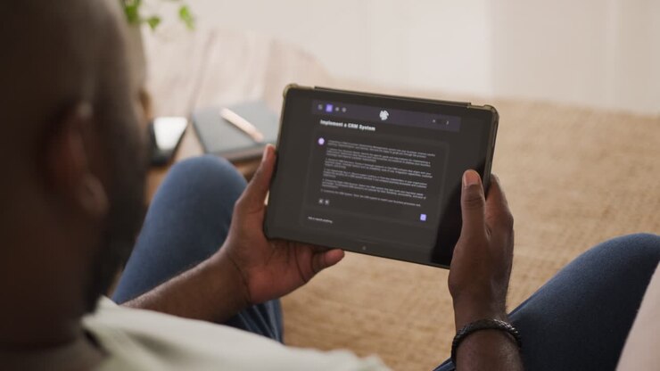 African American man sitting on couch holding tablet, adjusting grip to read dark settings screen
