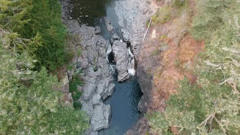 Downwards-panning-drone-shot-of-a-river-flowing-through-a-valley-that-gets-split-into-two-small-waterfalls-in-the-tropical-rainforest-of-British-Columbia,-Canada