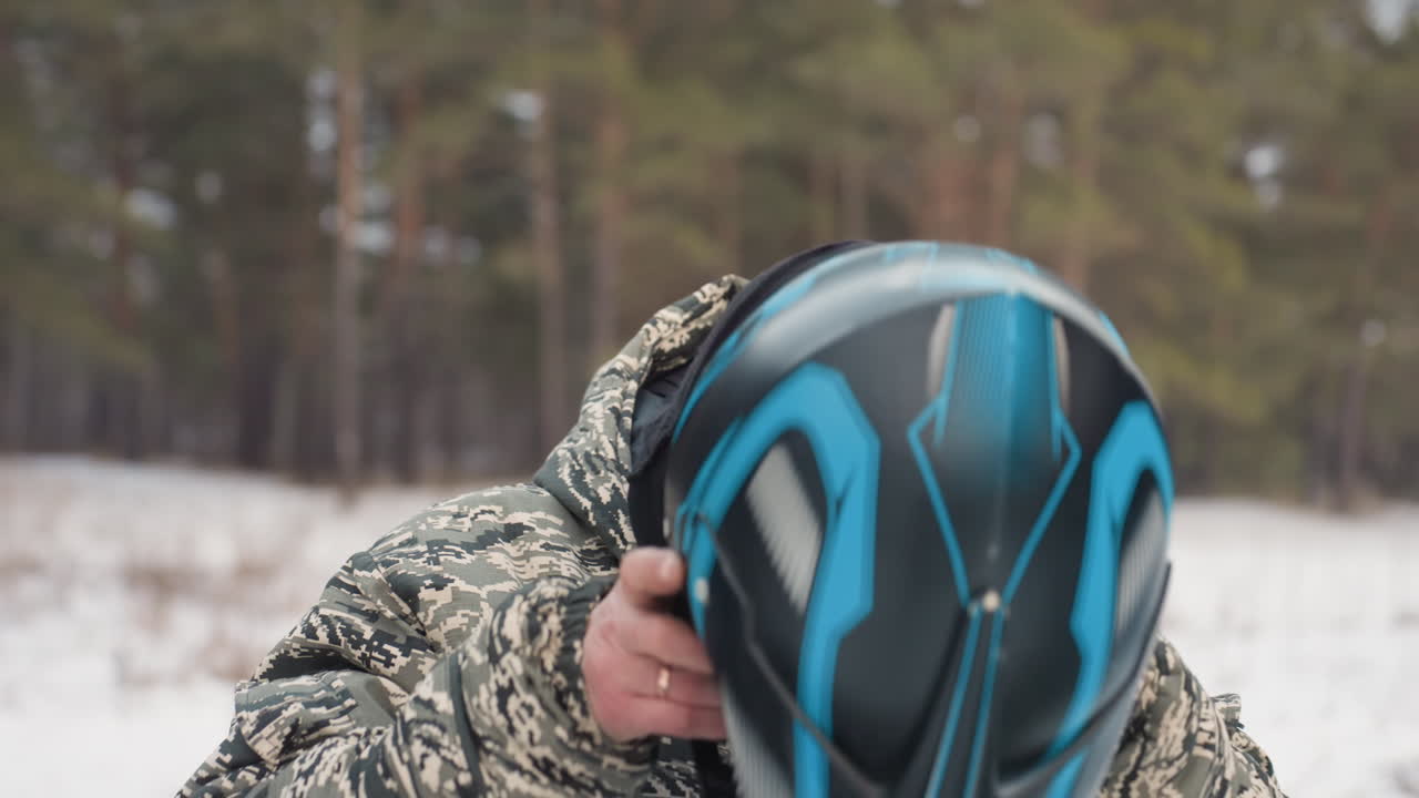 Un pilota uomo, in mimetica, prepara il casco con uno sfondo di pineta innevata. Ha il passamontagna alzato e sta regolando il cinturino con un'espressione concentrata. Un quad è parcheggiato nelle vicinanze e il suo respiro è visibile nell'aria fredda, indicando una prontezza che privilegia la sicurezza.