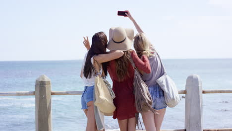 Three-young-woman-on-summer-vacation-taking-selfie-on-beach-promenade-wearing-denim-shorts