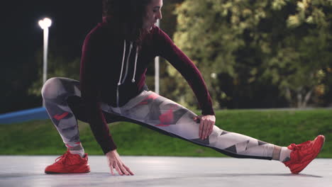 chica rizada deportiva entrenando y estirando las piernas en el parque por la noche