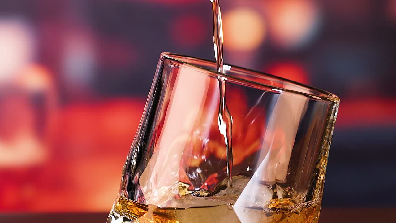 Un flusso di liquore dorato si versa in un bicchiere di cristallo pieno di cubetti di ghiaccio