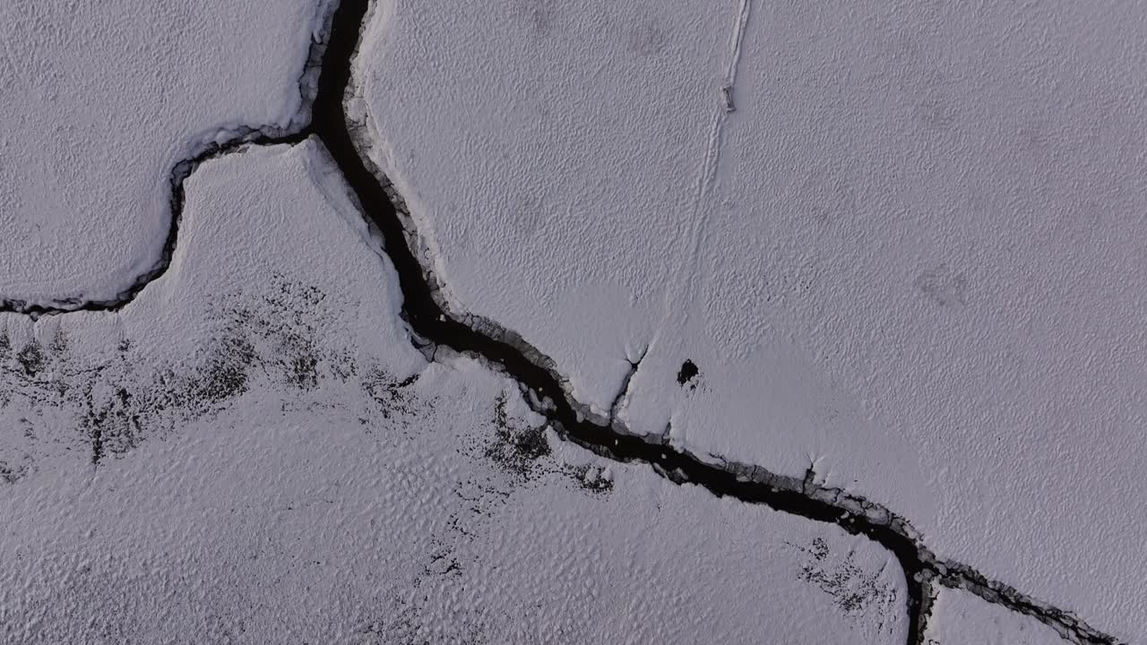 Top-down aerial view of a cracked frozen landscape in Borgarfjörður, Iceland, revealing a winding river channel cutting through snow-covered terrain in a stark winter setting.