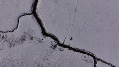top-down aerial view of a cracked frozen landscape in borgarfjörður, iceland, revealing a winding river channel cutting through snow-covered terrain in a stark winter setting.
