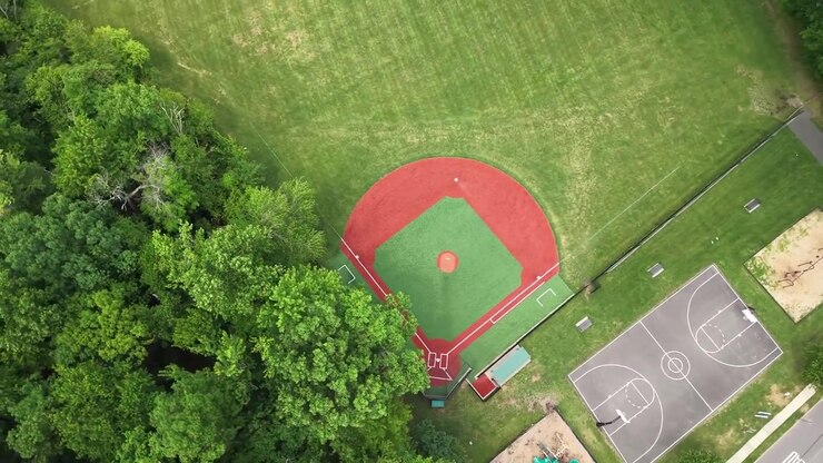 Ampia veduta aerea di un vivace parco verde con più campi da baseball, un parco giochi e un campo da basket, immerso tra alberi rigogliosi e un tranquillo quartiere residenziale sullo sfondo.