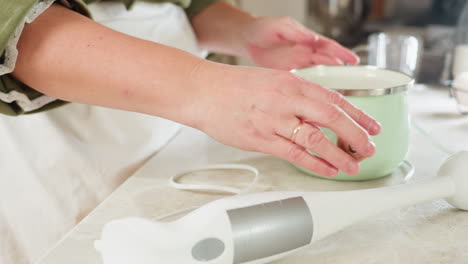 closeup hand view of woman in green sleeve holding light green pot with wooden handle while turning contents inside on kitchen counter, handheld blender visible nearby