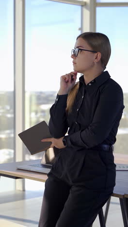pensive business woman in modern cabinet. young pretty woman in black clothes and glasses holding a pocketbook and thinks about new business project indoors. vertical video