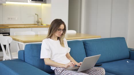 Blonde-woman-in-pajama-sit-on-the-couch-and-typing-on-silver-laptop