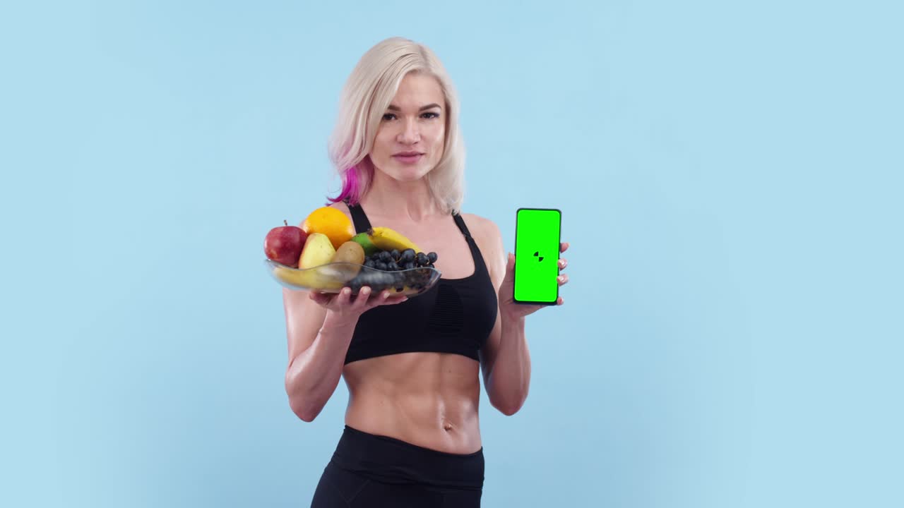 Beautiful woman with an athletic body holds plate of fruit and phone with an app, green screen. Muscular woman shows phone screen and plate of fruit in the studio, a blue background