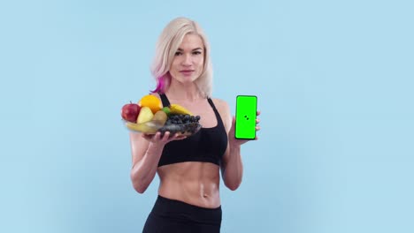 mujer hermosa con un cuerpo atlético sostiene un plato de fruta y teléfono con una aplicación, pantalla verde. mujer muscular muestra pantalla de teléfono y plato de frutas en el estudio, un fondo azul